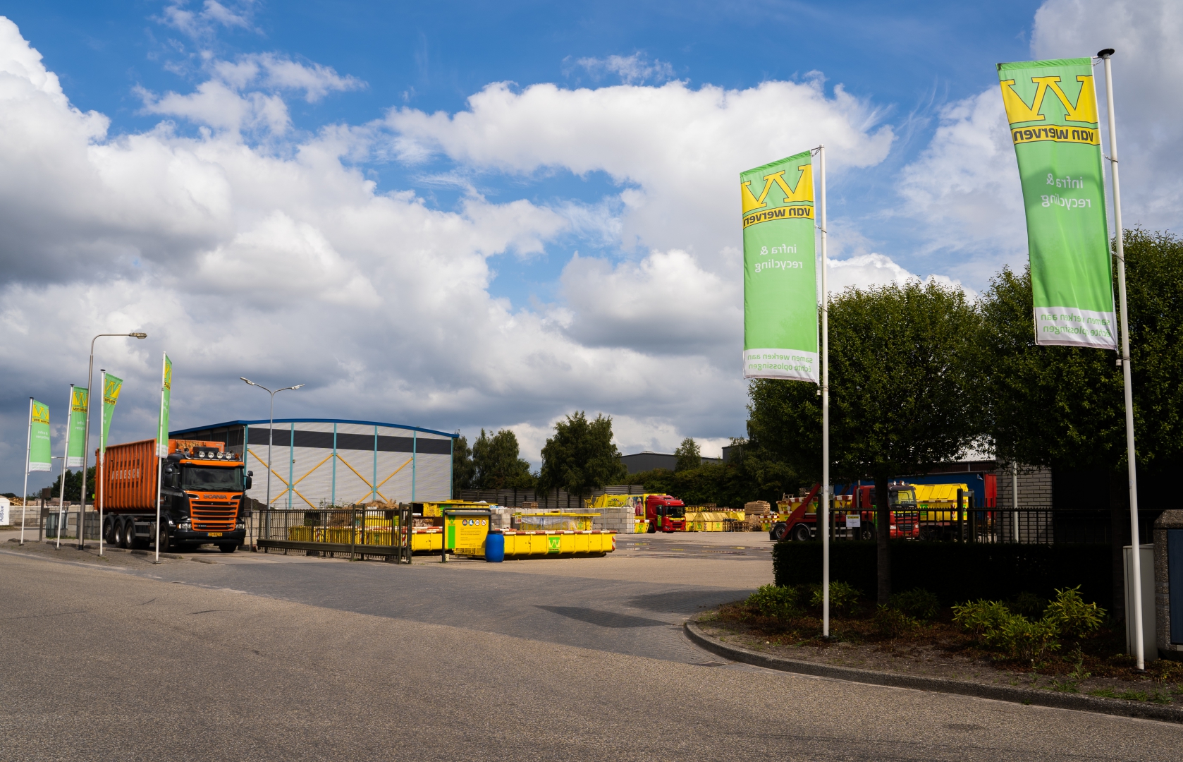 Ingang van de Van Werven-vestiging met groene bedrijfsvlaggen, een oranje vrachtwagen en gele containers voor de blauwe loods