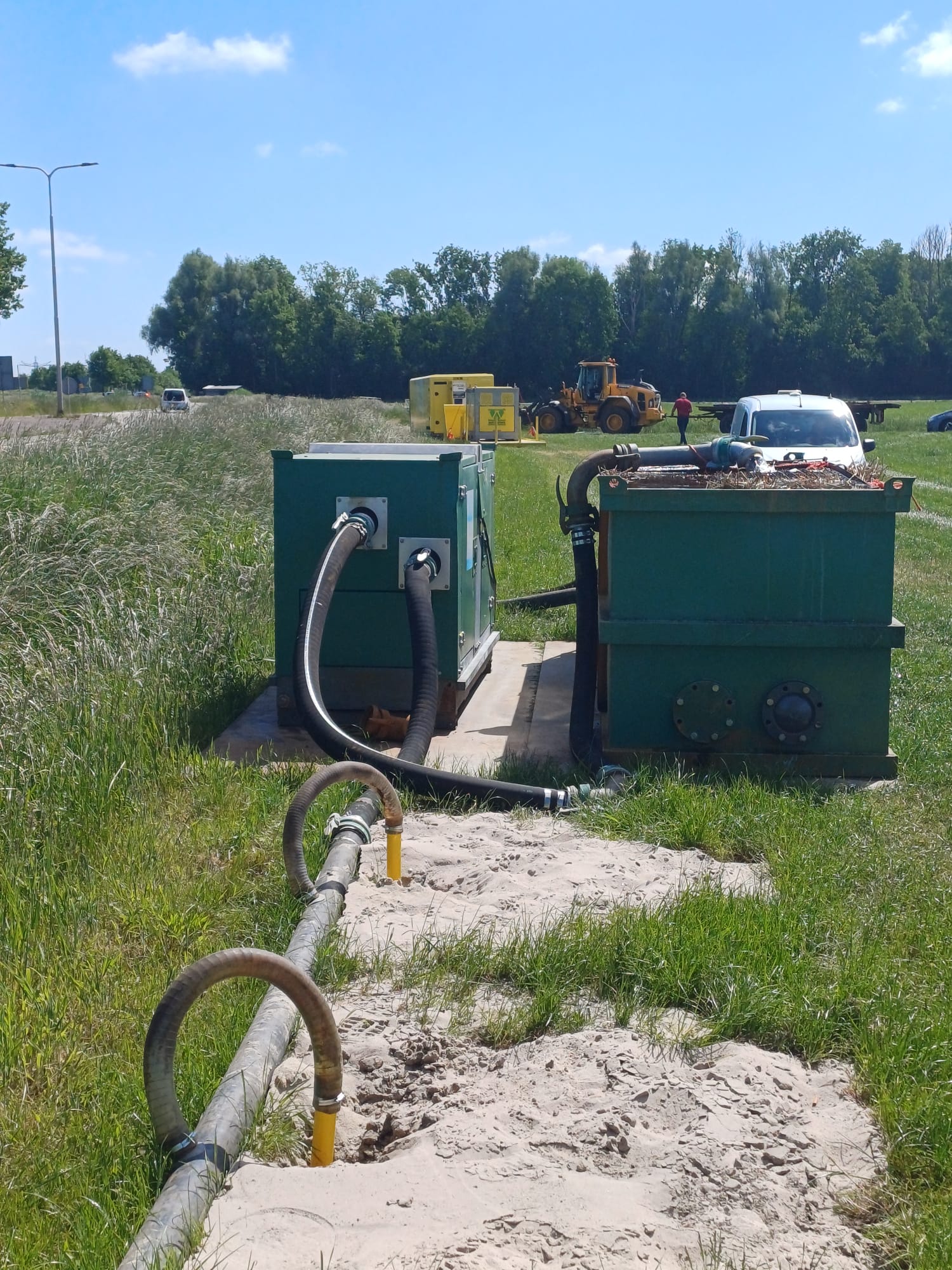 Twee donkergroene bemalingsunits in de berm langs een weg, met slangen over een zandpad naar de voorgrond.