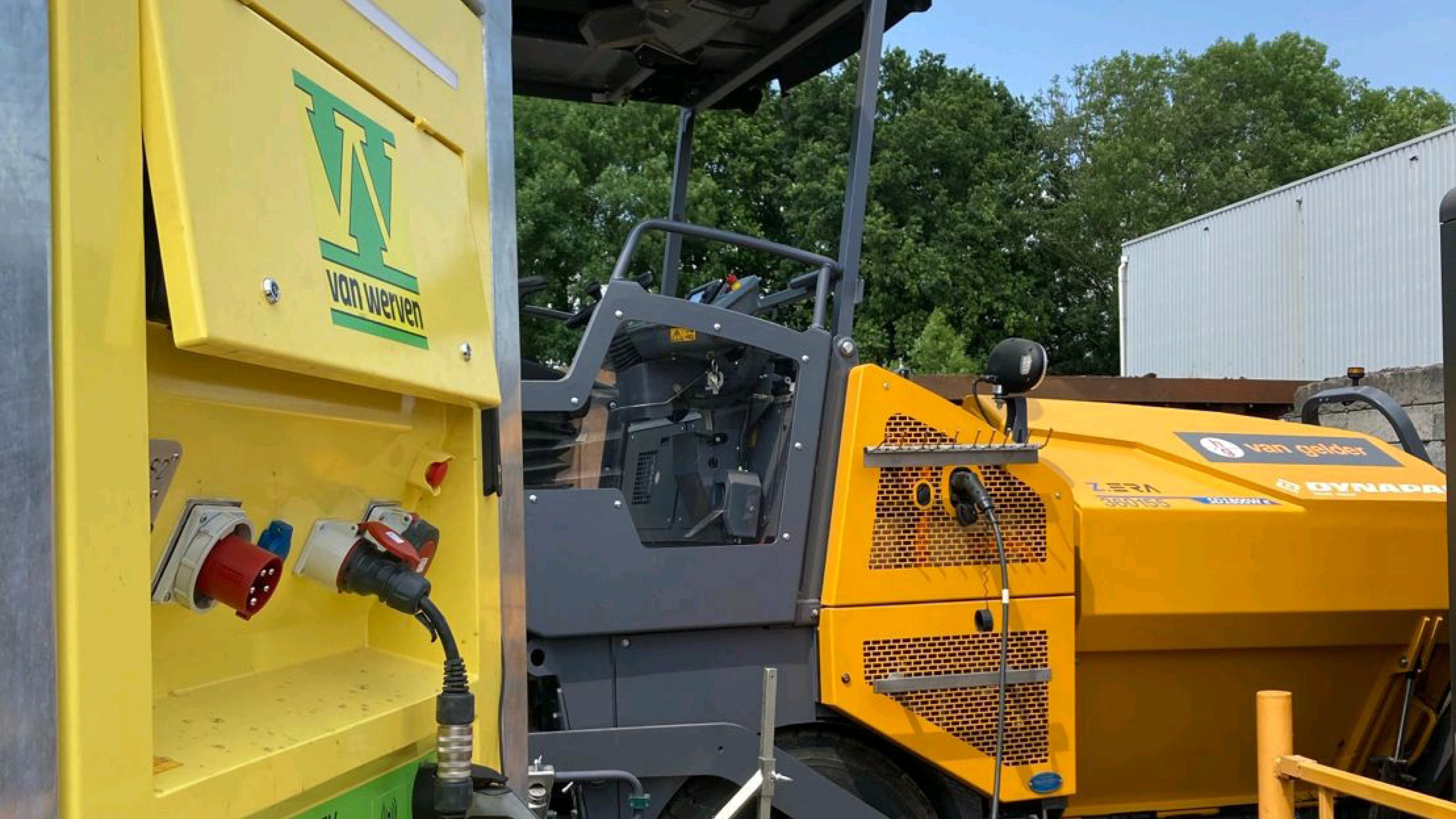 Een gele Van Werven-laadunit met stekkerdoos, aangesloten op een elektrische asfaltspreidmachine van Van Gelder.